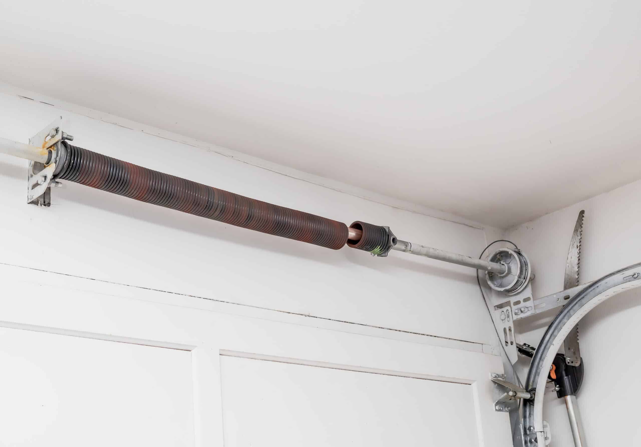 A broken garage door torsion spring mounted above the door with a gap in the coil indicating it has snapped. The metal hardware and tracks are visible on a white wall.