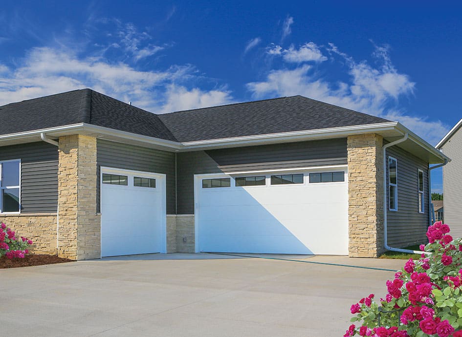 A contemporary house features two white garage doors with horizontal windows at the top. The exterior is a mix of stone and dark siding. Bright pink roses grow near the concrete driveway. The sky is clear with a few clouds.