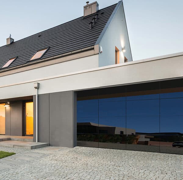 A modern house with a sleek design, featuring a pitched roof and large reflective garage door. The facade combines white and gray elements with minimalistic architecture. A stone path leads to the entrance, surrounded by a neatly trimmed lawn.