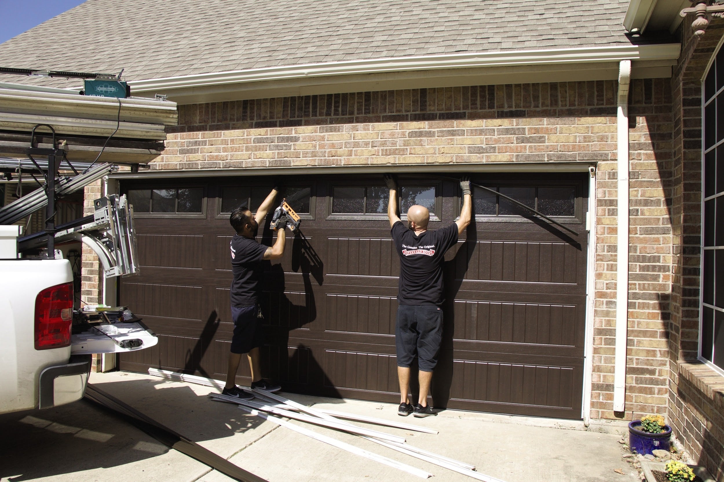 Two workers install a dark-colored garage door on a brick house. One stands on a ladder, while the other holds the door steady. A truck with equipment is parked nearby. The driveway is visible beneath them.