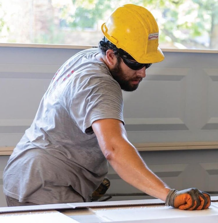 A man wearing a yellow hard hat, sunglasses, and gray shirt is working with tools. He is focused on his task, bending slightly forward. Behind him is a partially open garage door. Natural light is filtering in from outside.