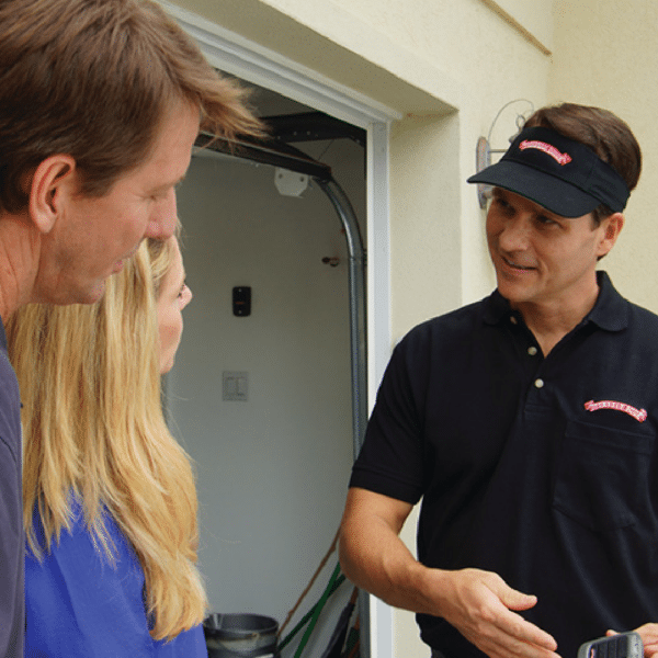 A person in a black polo and visor talks to two people, a man and a woman, near an open garage. The conversation appears amiable as they stand close to the garage entrance. The person holds a smartphone, showing something on it.