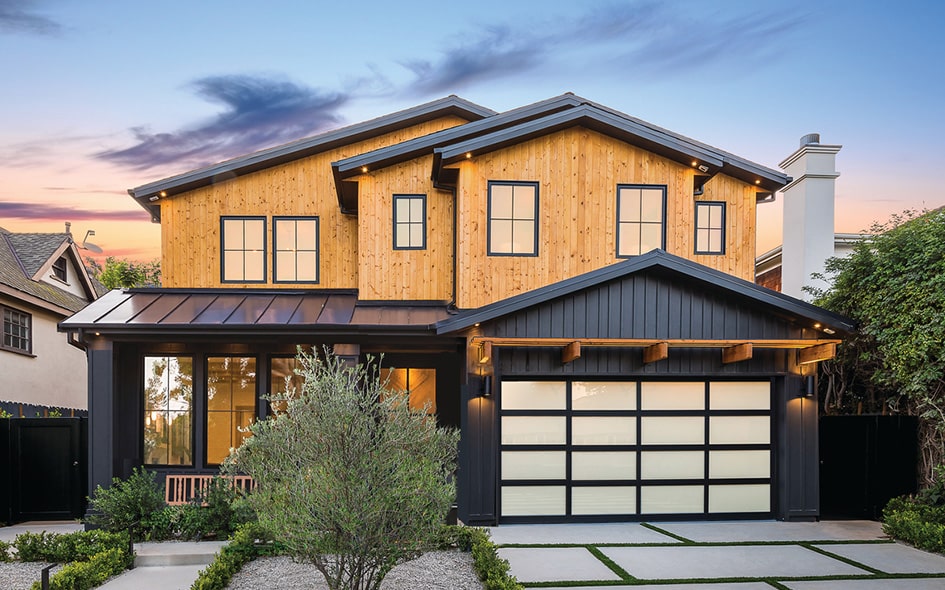Modern two-story house with a wooden facade and black trim. The ground floor features large glass garage doors. A small landscaped garden with shrubs and a stone pathway leads to the entrance. The sky is colorful at sunset.
