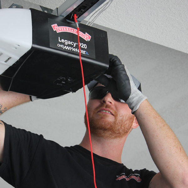 A person wearing gloves and a cap is inspecting or repairing a garage door opener attached to the ceiling. They are looking upwards and adjusting a component, with a red cord hanging down from the opener.