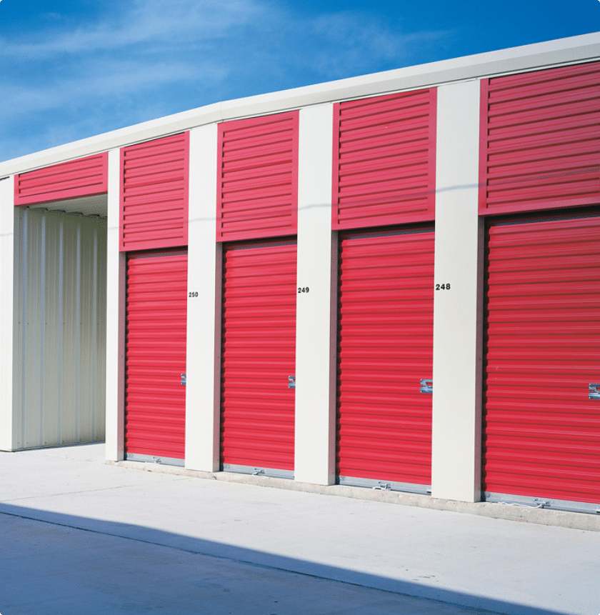 A row of outdoor storage units with red doors and white frames under a clear blue sky. Each unit door is closed, with numbers displayed on them. The ground is paved with concrete.
