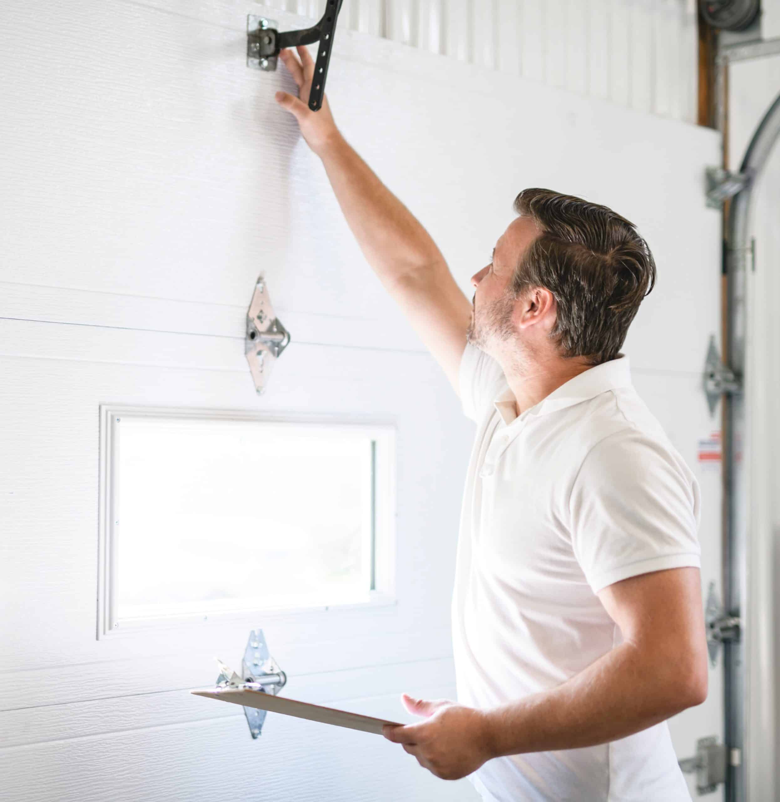 A man in a white polo shirt inspects a garage door mechanism, holding a clipboard in one hand and reaching up to touch the metal hardware above a window on the door.