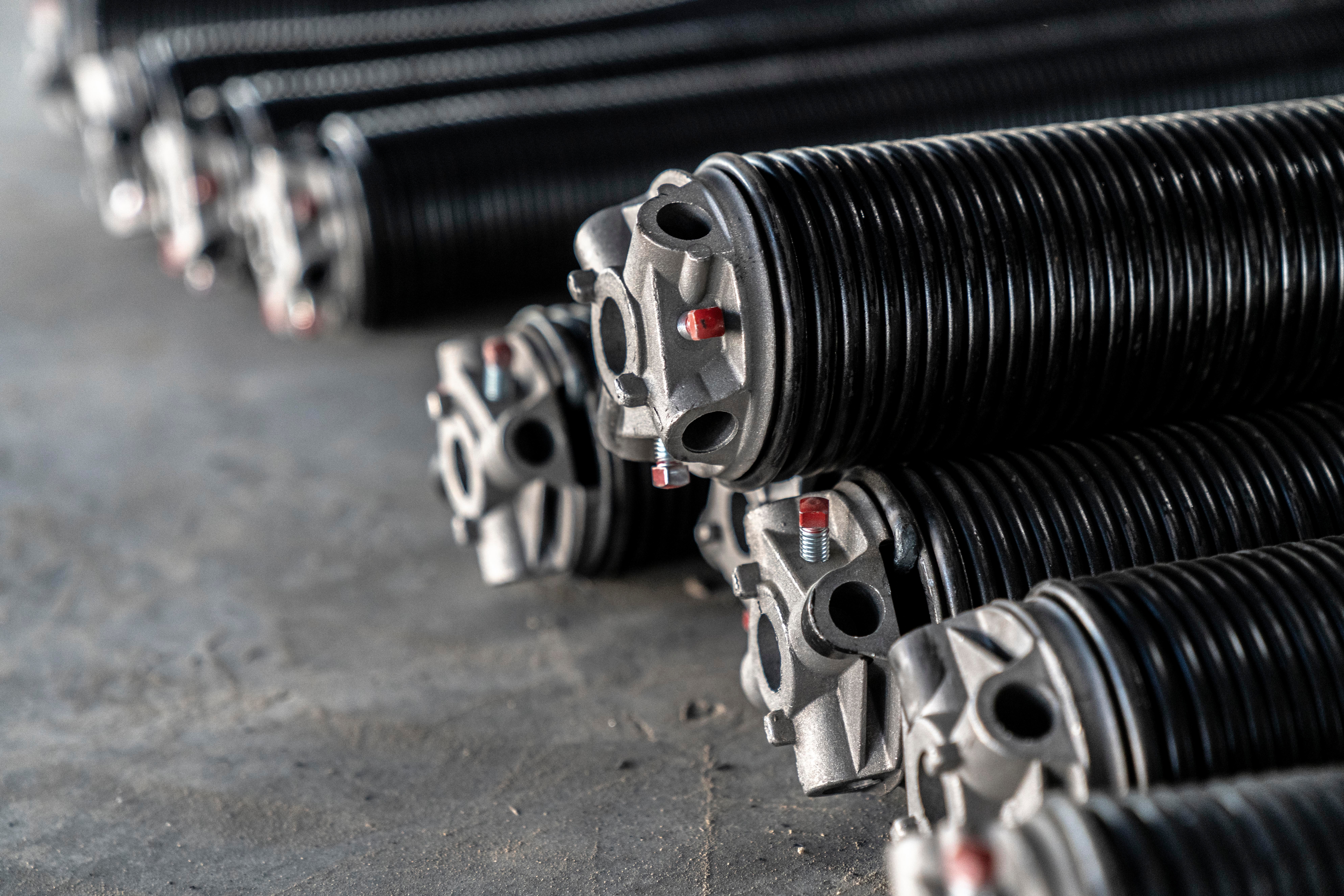 Close-up of several black coiled metal garage door springs with silver mounting ends arranged in parallel on a concrete floor.