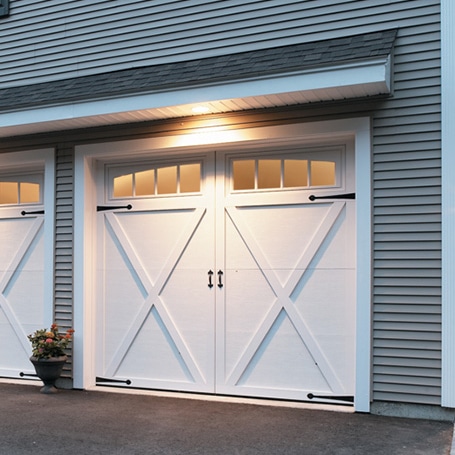 White double garage door with black hinges and handles, small top windows, gray siding, potted plant, and lit overhang nearby.