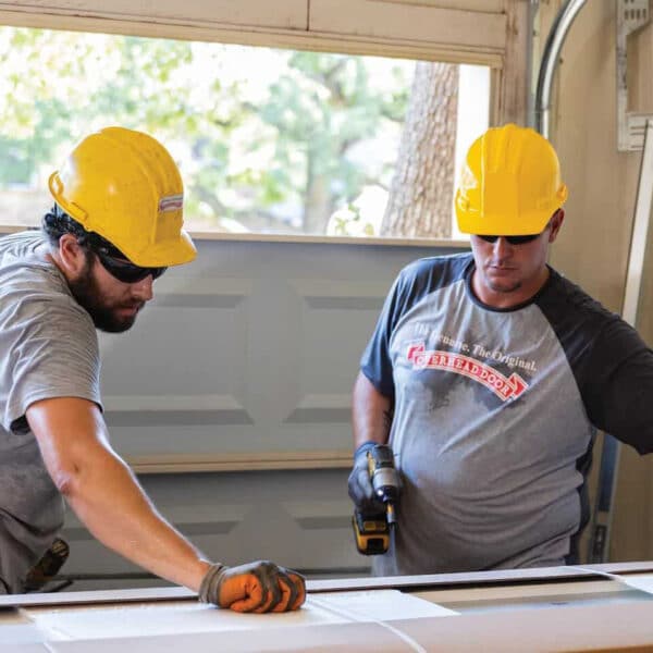 Two construction workers in yellow hard hats and gray shirts measure material indoors with tools as sunlight streams through a window.