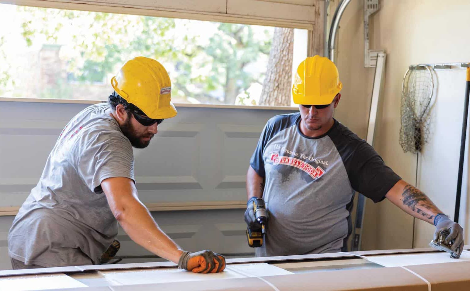 Two construction workers in yellow hard hats and gray shirts measure material indoors with tools as sunlight streams through a window.