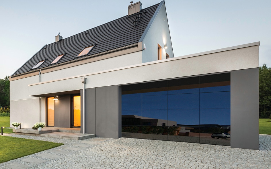 Modern house with grey and white exterior, black reflective garage door, sloped roof with skylights, lit entryway, cobblestone driveway.
