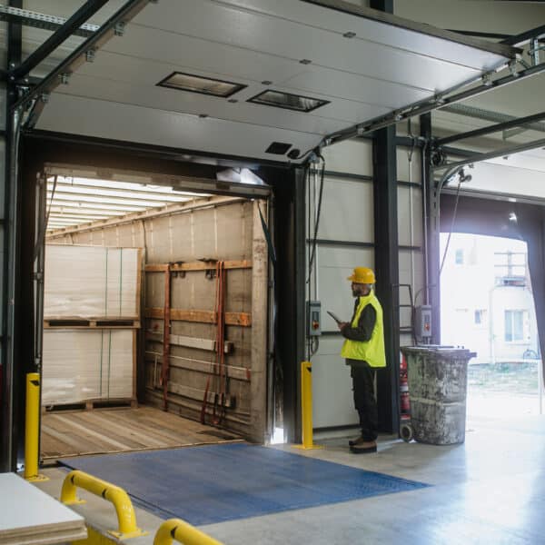 A worker in a yellow safety vest and hard hat stands by an open loading dock, using a tablet as pallets are unloaded from a truck.