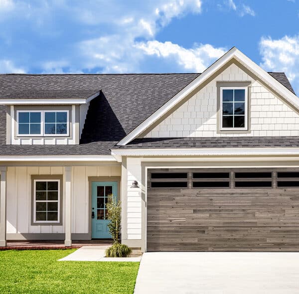 Modern suburban house with white siding, dark gray roof, large wooden garage door, and front porch under a bright blue sky.
