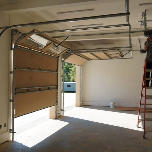 A person stands on a ladder repairing a garage door system inside a garage with two partially open doors. Sunlight streams in, and tools and equipment are scattered around.