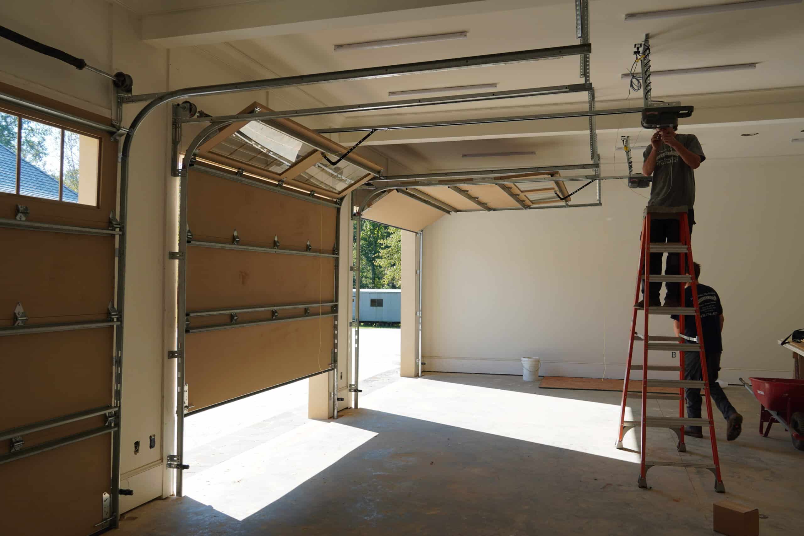A person stands on a ladder repairing a garage door system inside a garage with two partially open doors. Sunlight streams in, and tools and equipment are scattered around.
