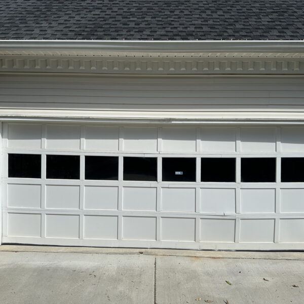 A white double-car garage door featuring rectangular windows along the top row is attached to a brick house. Potted plants are arranged on the left side near an exterior wall lamp.