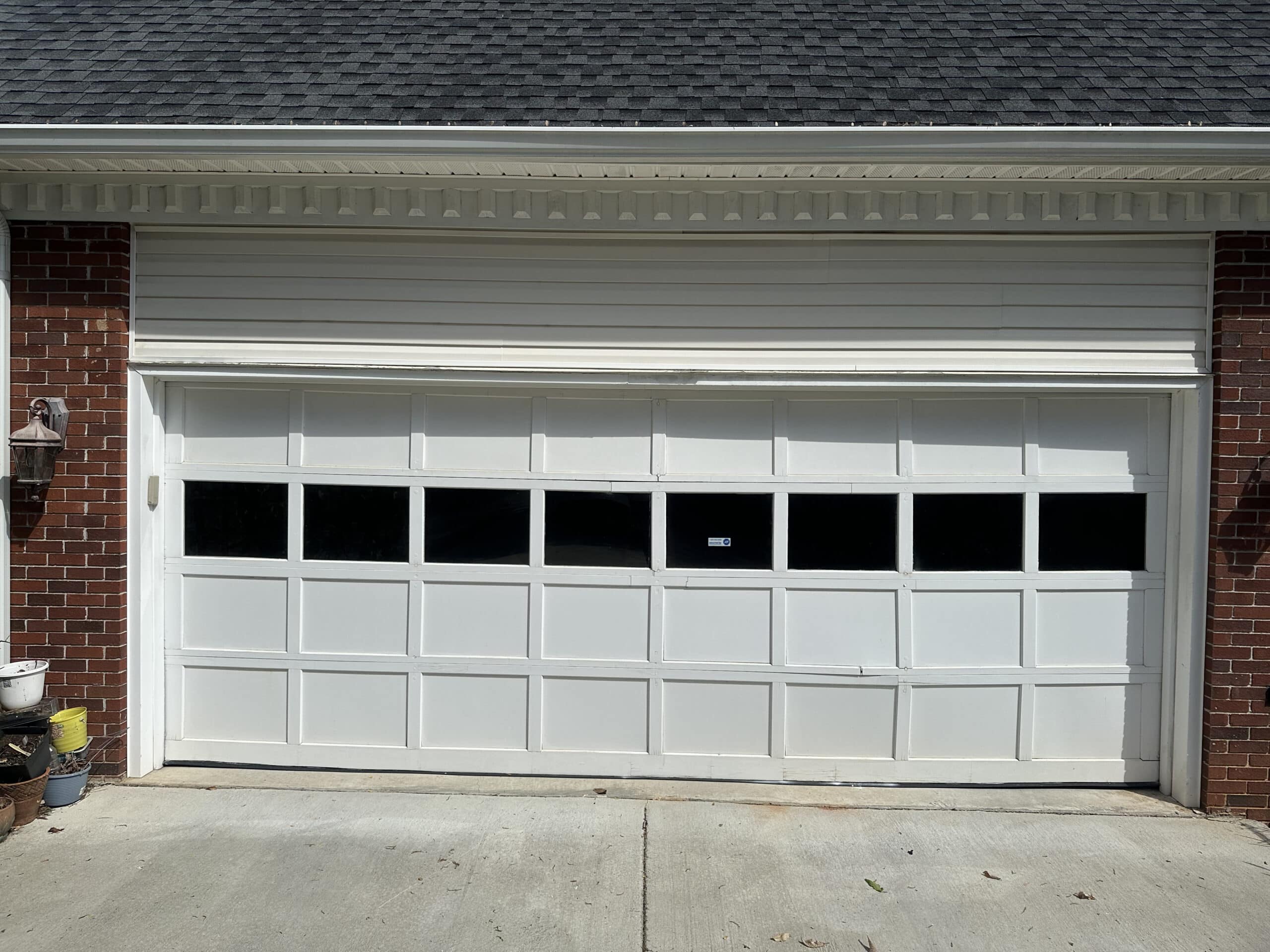 A white double-car garage door featuring rectangular windows along the top row is attached to a brick house. Potted plants are arranged on the left side near an exterior wall lamp.