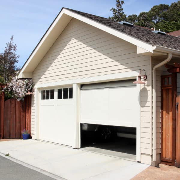 A cream-colored house featuring a partially open white garage door reveals a car inside. There’s a wooden fence, a potted plant beside the entrance, and trees in the sunny background.