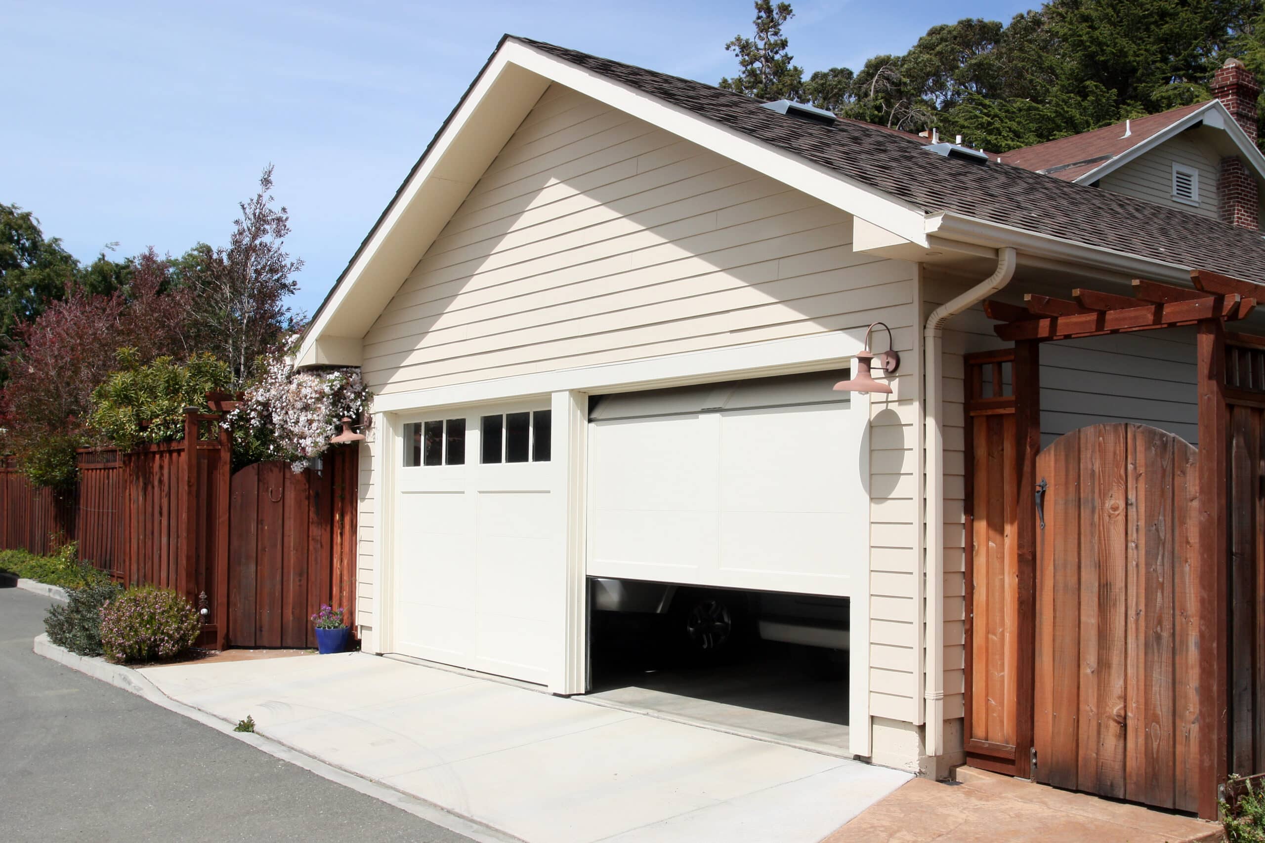 A cream-colored house featuring a partially open white garage door reveals a car inside. There’s a wooden fence, a potted plant beside the entrance, and trees in the sunny background.